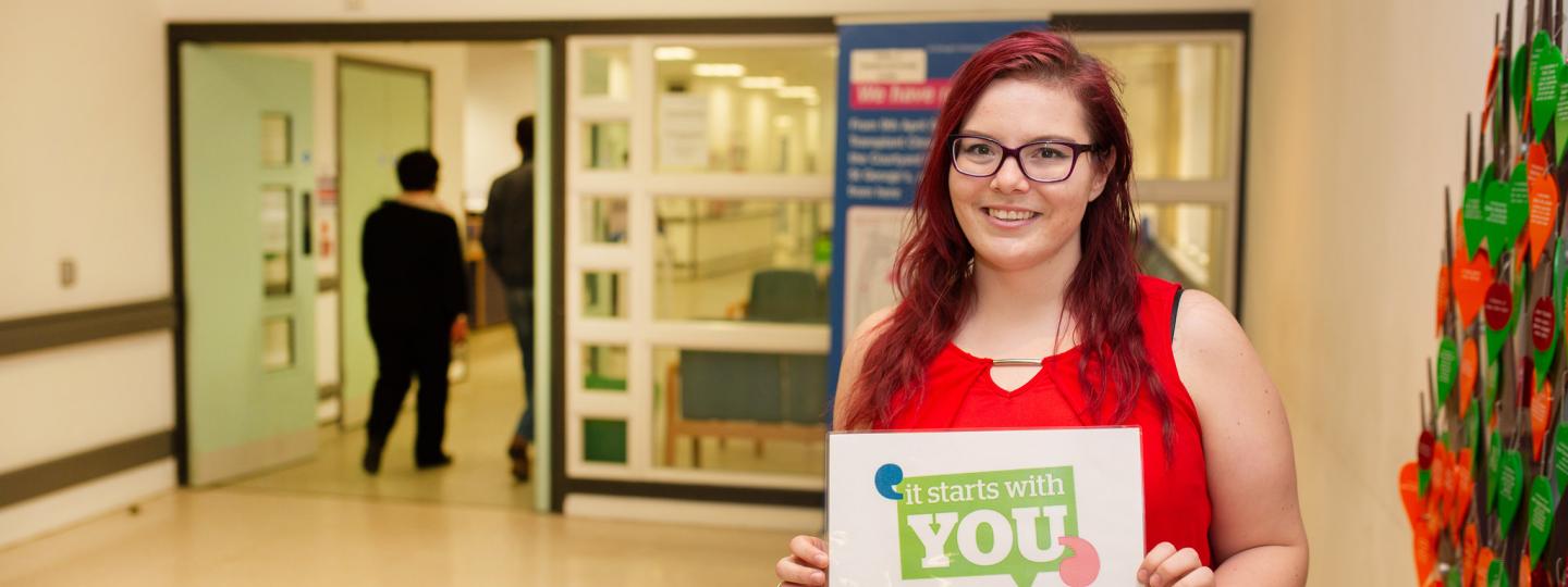 Woman stood in a hospital entrance holding a sign that says "It starts with you" and the Healthwatch logo